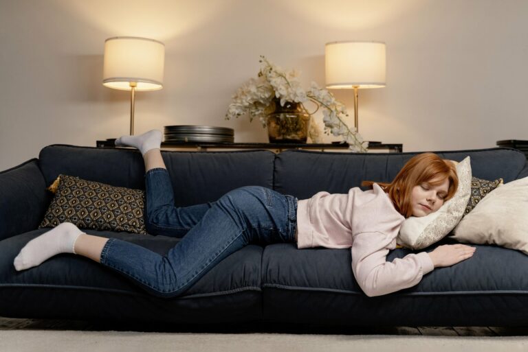 powernap.jpg Eine junge Frau mit roten Haaren liegt entspannt auf einem blauen Sofa, umgeben von Kissen und Lampen.