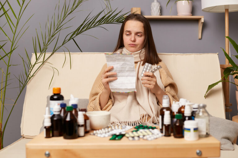 Eine kranke Frau sitzt auf einem Sofa, umgeben von verschiedenen Medikamenten und hält einen Zettel sowie eine Medikamentenpackung in der Hand.